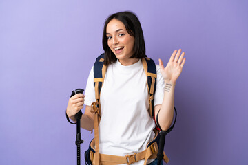young caucasian woman with backpack and trekking poles isolated on blue background saluting with han