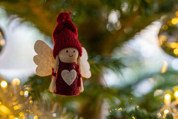 A red and white angel ornament hangs on a Christmas tree. The angel is wearing a red hat and has a heart on its chest. The tree is decorated with lights and the angel is the centerpiece of the tree