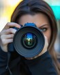 © Lakkhana - A female photographer peering through her camera lens, confident expression, natural lighting, blurred urban street background,