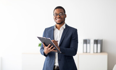 Happy young black male psychologist writing in clipboard, looking at camera and smiling in modern office. Psychotherapy services, mental health professional concept