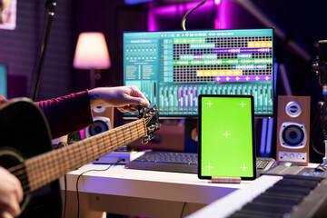 Fototapete - Artist songwriter practicing to play and tune his guitar by twisting knobs, watching online lesson via tablet with greenscreen display. Musician learning acoustic instrument skills in home studio.