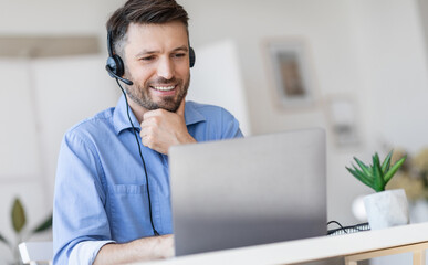 Hotline Support Service. Handsome male call center operator in headset working with laptop in modern office, looking at computer screen, free space