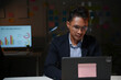 © amnaj - Young businessman working late at night in office using laptop, with graphs on a monitor in the background