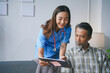 © amnaj - Young smiling nurse showing medical chart to senior patient sitting on couch at home, healthcare and medical concept