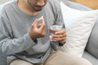 © KMPZZZ - Sick asian young man, male holding tablet pill on hand, painkiller medicine from fever, stomach pain, headache for treatment, take drug or vitamin with glass of water on at home. Health care concept.