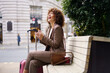 © Westend61 - Cheerful woman holding smart phone and sitting with suitcase at street