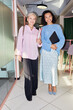 © Seventyfour - Vertical full length shot of senior and younger woman dressed in elegant smart casual outfits for office smiling at camera while standing in hallway of modern coworking space, camera flash
