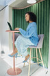 © Seventyfour - Full length shot of elegant multiethnic woman wearing skirt outfit for office in blue pastel colors sitting on comfy designer chair, working on laptop at standing desk in modern coworking space
