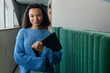 © Seventyfour - Portrait of young multiethnic woman dressed in comfy blue sweater smiling at camera with laptop in hands while posing in hallway at workplace, copy space