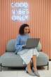 © Seventyfour - Vertical shot of cheerful multiethnic woman in smart casual outfit browsing on laptop while working on comfy seat in lounge area of modern coworking space