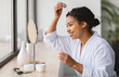 © Prostock-studio - African American woman smiles as she applies serum to her hair while looking in a vanity mirror. The setting is a well-lit, contemporary bathroom with stylish decor.