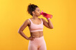 © Prostock-studio - A young woman wearing workout attire stands against a bright yellow backdrop. She drinks from a pink sports bottle, taking a moment to hydrate during her fitness routine.