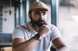 © BullRun - Serious caucasian mature man visitor of cafe sitting near window pondering on ideas and future plans, serious bearded owner of small business in hat and with trendy wrist watch concentrated