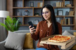 © Liubomir - Asian woman holding smartphone and pizza, smiling at home. Engages in social media or online activity, enjoying casual relaxation and comfort with delicious takeaway meal.