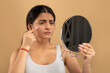 © Prostock-studio - In a warm-toned indoor space, a young Indian woman focuses on her skincare routine, using a mirror to assess her skin. She is thoughtfully engaged in self-care activities.