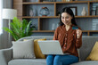 © Liubomir - Asian woman shopping online using laptop and credit card, sitting comfortably on sofa in home setting. Concept of convenience, technology, and e-commerce in everyday life.