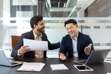 Two cheerful professionals are sitting at a modern office table, engaged a lively conversation. The environment suggests a positive collaboration, with documents, laptops, indicating work activities