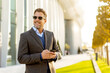 © BGStock72 - Confident businessman enjoys a sunny afternoon while checking his phone near a modern building