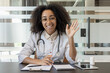 © Tetiana - Portrait of a young African-American female doctor sitting at a desk in a hospital, talking to the camera via online communication, greeting and waving
