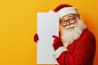 © Maxim Borbut - Santa Claus, appearing happy, looks through an open space behind a blank sign on a yellow backdrop, leaving room for copy.