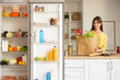 © Pixel-Shot - Young woman with paper bag of food on counter near open fridge in kitchen