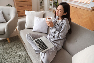 Sticker - Young woman in grey pajamas with coffee and laptop working on sofa at home