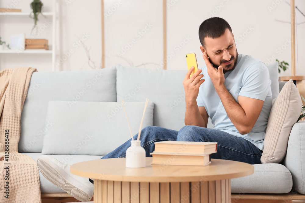 Young man with mobile phone suffering from ear pain on sofa at home