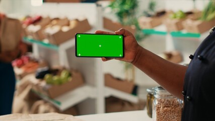 African american owner using greenscreen display at checkout, holding isolated mockup template while trying to sell organic produce at local shop. Merchant showing chromakey layout.