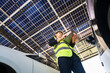 © ultramansk - Engineer inspecting solar panels in a solar-powered carport surrounded by parked cars. Renewable energy, clean technology, sustainable infrastructure, and solar panel performance monitoring.