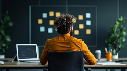 Leinwandbilder - focused man in yellow shirt sits at desk, brainstorming ideas in office. laptop and coffee cup are present, with sticky notes on wall
