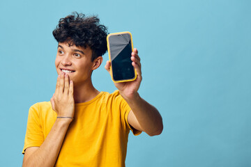 Wall Mural - Young man smiling while holding a smartphone against a blue background, wearing a bright yellow shirt, showcasing modern technology in a cheerful and engaging manner