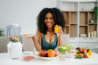 © Nuttapong punna - Smiling woman in a modern kitchen with fresh fruits, vegetables, blender, and orange juice, promoting clean eating, wellness