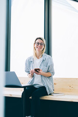 Wall Mural - happy hipster girl in trendy apparel and glasses smiling at camera and posing during freelance job i