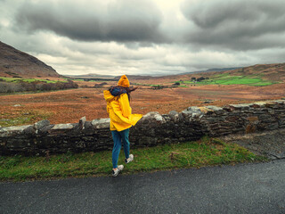 Wall Mural - Teenager girl tourist in yellow hat and jacket looking from a hill at stunning nature scenery with a valley, mountains and dramatic sky. Travel and sightseeing in Connemara, Ireland.