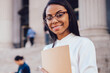 © BullRun - Portrait of prosperous cheerful african american student dressed in white shirt smiling at camera while updating profile in networks on smartphone.Positive dark skinned female lawyer with folder