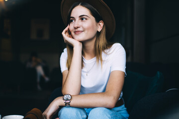 Smiling caucasian hipster girl in trendy hat and casual clothes sitting indoors enjoying free time, positive 20s young woman dressed in stylish apparel looking away dreaming about vacations