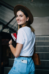 Smiling beautiful caucasian hipster girl in trendy hat walking on stair indoors looking at camera, portrait charming young teenage female dressed in stylish outfit carrying book for studying