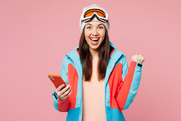 Wall Mural - Skier young woman wear warm blue padded jacket ski goggles mask spend weekend winter season in mountains hold use mobile cell phone do winner gesture isolated on plain pink background. Hobby concept.