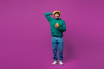 Wall Mural - Full body young surprised Indian man he wear green hoody yellow hat casual clothes put hand on head hold use mobile cell phone isolated on plain purple background studio portrait. Lifestyle concept.