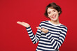© ViDi Studio - Young smiling happy Caucasian woman she wearing blue striped sweatshirt casual clothes point hands arms aside on empty area mockup isolated on plain red background studio portrait. Lifestyle concept.
