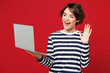 © ViDi Studio - Young happy IT woman she wear blue striped sweatshirt casual clothes hold use work on laptop pc computer leading video conference call waving hand isolated on plain red background. Lifestyle concept.