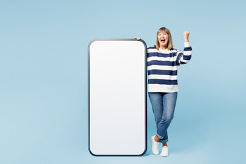 Poster - Full body elderly blonde woman 50s years old wears striped sweatshirt casual clothes big huge blank screen mobile cell phone smartphone with area do winner gesture isolated on plain blue background.