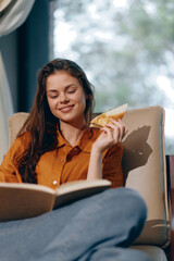 Canvas Print - A happy young woman enjoying a healthy snack while reading a book in a cozy indoor setting, wearing a stylish amber shirt and relaxed denim