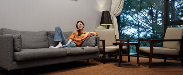 Wall Mural - Young woman relaxing on a sofa in a cozy living room with natural light, wearing casual orange top and jeans, conveying comfort and tranquility