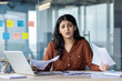 © Liubomir - Businesswoman sitting at her desk while looking stressed amidst paperwork and a laptop.