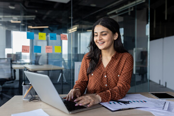 Wall Mural - Young professional woman typing on a laptop, analyzing data charts, and brainstorming ideas in an office.