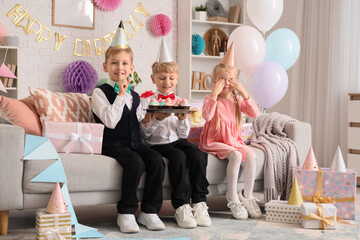 Wall Mural - Cute little happy kids in party hats with cupcakes and gift boxes showing silent gesture while celebrating birthday at home