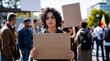 © Sachin - Young Mediterranean woman with curly dark hair holding environmental protest sign at climate change rally, serious expression, crowd with placards in background