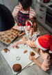 © Soloviova Liudmyla - Cute little girls in red Santa hats with mother making homemade dough Christmas gingerbread cookies using cookie cutters together in home kitchen. Happy family holidays preparation, childhood concept.