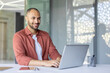 © Liubomir - A professional man using a laptop while seated at a desk in a bright, modern workspace, exuding positivity and focus. The scene highlights productivity, business, and a pleasant working atmosphere.